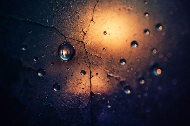Macro photo of cracked glass with layered textures beneath, water droplets refracting tiny cityscapes, all lit by warm, cinematic backlight.